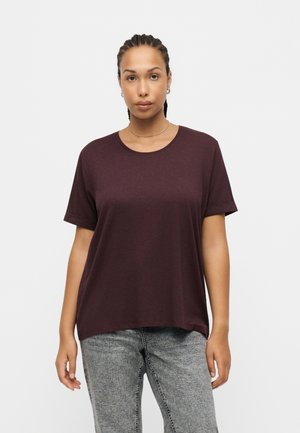 Person med flettet hår har en løs bordeaux t-shirt og grå jeans på, står mod en ensfarvet hvid baggrund og vender fremad.