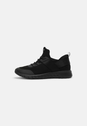 Baskets de sport noires avec tige en maille, sangle élastique, semelle rembourrée et tirette, sur fond blanc, vue de côté.