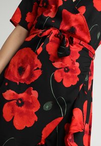 Černé látkové šaty s překládaným designem a velkými červenými mákovými vzory. Součástí je odpovídající látkový pásek a krátké rukávy.