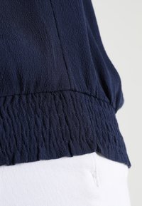 Marinblå strukturerad topp med en draperad nederkant, tillverkad av ett lätt material, kombinerad med vita jeans eller byxor.