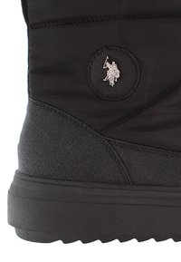 Bota aislante negra con un tejido texturizado, suela de goma y un parche redondo con un emblema plateado de un jinete.