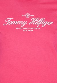 Lyserød bomuld t-shirt med hvid trykt tekst: "Tommy Hilfiger," "EST. 1985," og "REGISTERED TRADEMARK NEW YORK." Glat tekstur, rund halsudskæring.