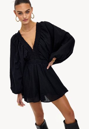 Robe noire en tissu texturé, décolleté en V profond, manches larges et taille froncée. Elle présente une coupe décontractée et s'arrête au-dessus de la mi-cuisse.