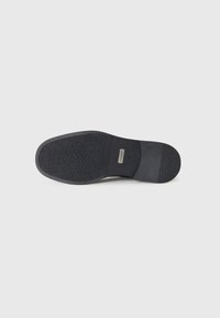 Svart gummisåle med strukturert grep mønster, glatte områder og en sølvmerke nær hælen. Ergonomisk form for stabilitet.