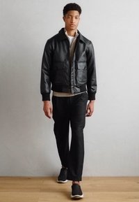 Veste bomber en cuir noir avec deux poches avant, chemise superposée grise en dessous, pantalon noir et chaussures à enfiler. Design minimaliste.