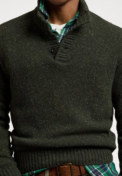 Maglione lavorato a maglia verde scuro con colletto alto e due bottoni, caratterizzato da un tessuto testurizzato e polsini a coste. Una camicia a quadri sbircia sotto.