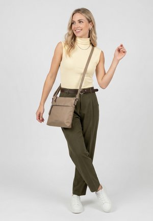 Lächelnde Frau trägt ärmellosen gelben Rollkragenpullover, olivgrüne Hose, weiße Sneaker, braunen Gürtel und eine beige Umhängetasche vor hellgrauem Hintergrund.