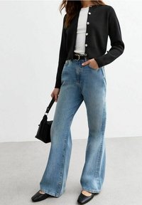 Schwarze Strickjacke mit silbernen Knöpfen, weißes Oberteil, hochtaillierte hellblaue ausgestellte Jeans, schwarze Ballerinas und eine kleine schwarze Handtasche.