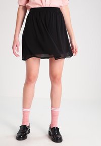 Kvinna som bär en svart kjol med elastisk midja, rosa skjorta, rosa randiga strumpor och svarta loafers och står framför en ljus enfärgad bakgrund.