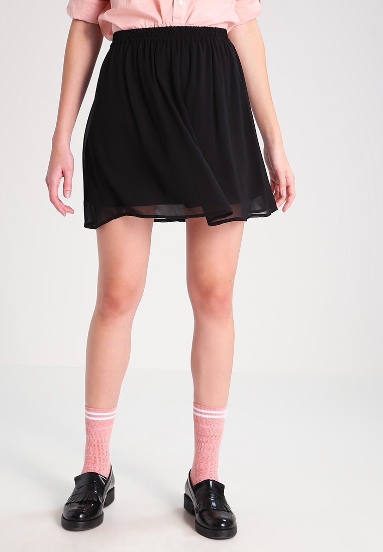 Kvinna som bär en svart kjol med elastisk midja, rosa skjorta, rosa randiga strumpor och svarta loafers och står framför en ljus enfärgad bakgrund.