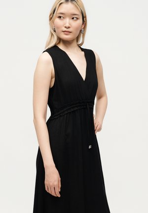 Młoda kobieta o jasnych blond włosach, ubrana w czarną sukienkę bez rękawów z głębokim dekoltem w serek i talią podkreśloną paskiem, patrzy na bok na jednolitym tle.