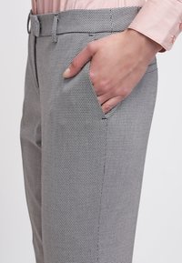 Main dans la poche droite d'un pantalon gris à carreaux porté avec une chemise rose clair à manches longues.