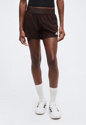 Person iført sorte sportsshorts, hvide mellemlange sokker og hvide sneakers med sorte striber, stående mod en neutral baggrund.