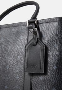 Borsa tote in pelle nera testurizzata con manico liscio. Presenta un'etichetta per bagagli e un motivo logo sottile in una tonalità più scura.