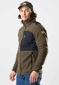 Fleecejacke in Olivgrün mit schwarzen Einsätzen. Verfügt über eine mit Reißverschluss verschließbare Brusttasche, Seitentaschen und strukturierten Stoff. Kombinierbar mit blauen Jeans.