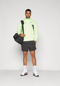 Veste coupe-vent vert fluo, accents noirs, short noir, baskets blanches avec bord rouge, portant un sac noir, debout contre un fond gris.