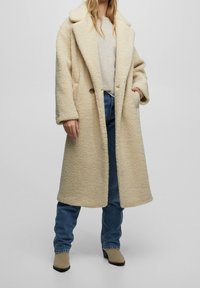 Manteau beige texturé oversize avec revers crantés, deux boutons et poches latérales, porté sur un pull léger et un jean bleu.
