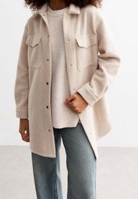 Chaqueta de forro polar beige claro con dos bolsillos en el pecho, botones a presión y un ajuste relajado, llevada sobre un suéter de punto ligero y jeans azules.