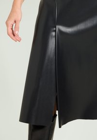 Sort lædernederdel med sidelommer, glat tekstur og et elegant design. Synlig hånd på stoffet for skala.