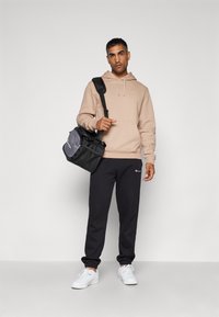 Beige Hoodie mit vorderer Kängurutasche, schwarze Jogginghosen und weiße Sneaker. Das Modell hält eine grau-schwarze Umhängetasche. Einfacher Hintergrund.