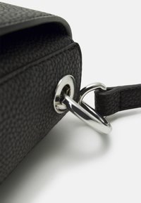 Angolo di una borsa in pelle nera testurizzata con hardware in metallo argentato e attacco ad anello. La tracolla è anch'essa nera e presenta una finitura liscia.