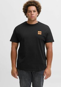 Schwarzes Baumwoll-T-Shirt mit kurzen Ärmeln und einem quadratischen orangefarbenen Logo auf der linken Brust. Getragen mit dunklen Jeans, steht das Modell vor einem einfarbigen Hintergrund.