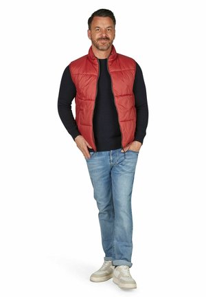 Mand stående med hænderne i lommerne iført rød quiltet vest, sort langærmet skjorte, lyseblå jeans og hvide sneakers på hvid baggrund.