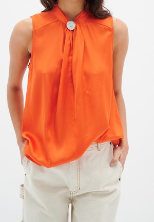 Vrouw draagt mouwloze feloranje blouse met een zilveren knoop bij de hals, gecombineerd met offwhite broek, handen in de zakken.