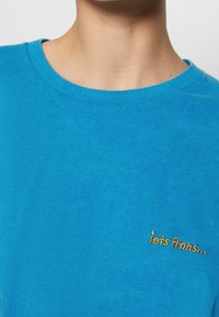 Turquoise cotton T-shirt with a round neckline, featuring a small golden embroidered text "iets frains..." on the left side.