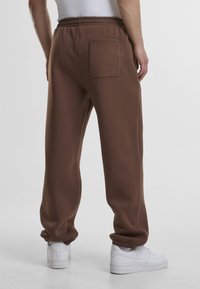 Pantalon de survêtement marron avec une coupe décontractée, une taille élastique, des poches latérales et une seule poche arrière. Le tissu semble doux et texturé.