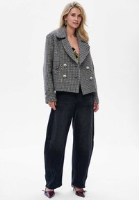 Blonde vrouw in een grijze houndstooth dubbelbreasted jas, zwarte wijde broek en bordeauxrood puntige hakken op een witte achtergrond.