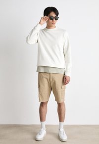 Ανοιχτό κρεμ φούτερ φορεμένο πάνω από μια θαμπή πράσινη μπλούζα, συνδυασμένο με μπεζ cargo σορτς. Λευκά αθλητικά παπούτσια και σκούρα γυαλιά ηλίου ολοκληρώνουν την εμφάνιση.