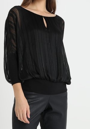 Femme portant un chemisier noir transparent rayé avec un col en trou de serrure et des poignets ajustés, associé à un pantalon noir sur un fond clair.