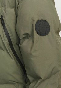 Zelena podstavljena jakna s teksturiranim materijalom, s crnim zatvaranjem na patentni zatvarač i kružnom crnom logotipskom oznakom na rukavu.