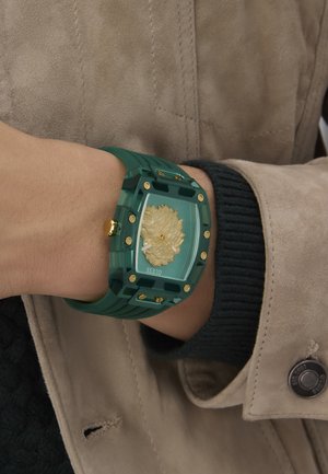 Grüne Gummi-Uhr mit klarem Zifferblatt, das ein goldenes Löwenmotiv zeigt, goldenen Hardware-Akzenten und einem wuchtigen rechteckigen Design.