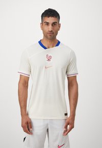 Maglia da calcio bianca con colletto blu, caratterizzata da un logo di gallo e l'emblema 'FFF'. Logo Nike in corallo sul petto, tessuto testurizzato.