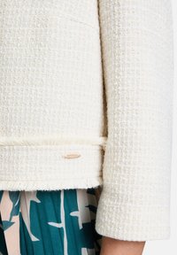 Jasno-beżowa kurtka o fakturowej strukturze z plecionym wzorem, postrzępionym dołem i małą etykietą w pobliżu dolnej krawędzi. Materiał ma lekko wypukłą powierzchnię.