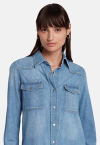 Camicia in denim azzurro chiaro con bottoni frontali, colletto a punta e due tasche sul petto. Presenta bottoni in metallo argentato e una finitura leggermente strutturata.