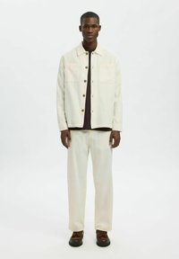 Giacca a bottoni leggera color crema con due tasche sul petto, abbinata a pantaloni coordinati. Una camicia marrone sotto e scarpe con lacci marroni completano il look.
