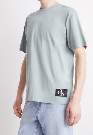 Mężczyzna noszący jasnoszary t-shirt z krótkim rękawem z naszywką z logo Calvin Klein Jeans przy krawędzi, w połączeniu z jasnoniebieskimi dżinsami.