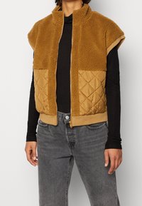 Beżowa kamizelka zapinana na zamek, z miękkim polarem na górze i pikowanym materiałem na dole, z dwoma przednimi kieszeniami, wysokim kołnierzem i ściągaczem u dołu.