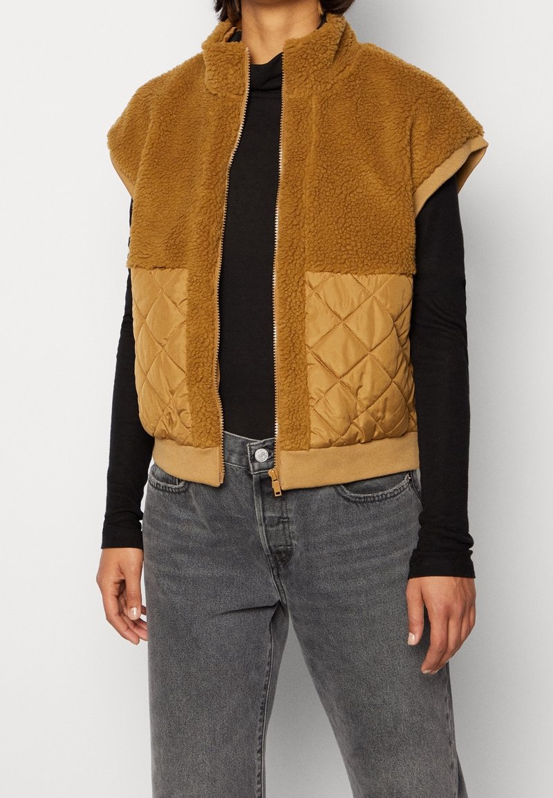 Beżowa kamizelka zapinana na zamek, z miękkim polarem na górze i pikowanym materiałem na dole, z dwoma przednimi kieszeniami, wysokim kołnierzem i ściągaczem u dołu.