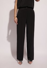 Czarne, wysokie spodnie z dopasowanym krojem, z wyraźnym szwem z przodu, okrągłym kółkiem na pasek i gładką teksturą. Srebrne obcasy dopełniają stylizację.