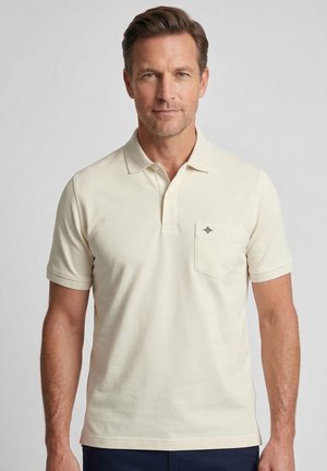 Homme portant un polo beige à manches courtes avec une poche poitrine et un petit logo, debout devant un fond clair uni.