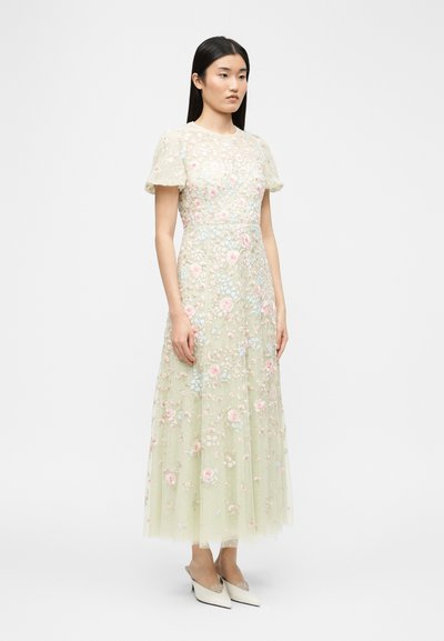 Μια γυναίκα στέκεται φορώντας ένα ανοιχτοπράσινο, κοντομάνικο, φόρεμα μέχρι τον αστράγαλο με παστέλ κεντήματα λουλουδιών και λευκά ψηλοτάκουνα παπούτσια με μυτερή μύτη.
