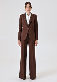Bruin maatpak met een getailleerde jas, brede broekspijpen en een witte blouse. Zilveren hardwaredetails op de jas. Gladde textuur.