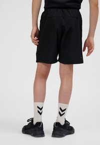 Pantaloncini sportivi neri con vita elastica, lunghezza sopra il ginocchio, abbinati a calzini color crema con motivi a chevron neri e scarpe nere.