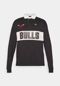 Črna polo srajca dolgimi rokavi s belim horizontalnim trakom, velikim napisom "BULLS", vezenim logotipom bika in kontrastnim ovratnikom. Narejena iz bombaža.