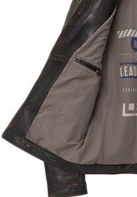 Binnenkant van een leren jas, donkerbruin leren accent, grijze stofvoering, bevat een ritszak, gedrukt logo en tekstdetails.