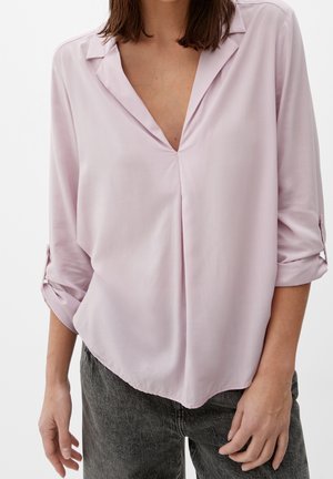 Femme portant un chemisier rose clair à manches longues avec col en V et manches retroussées, associé à un jean gris foncé.